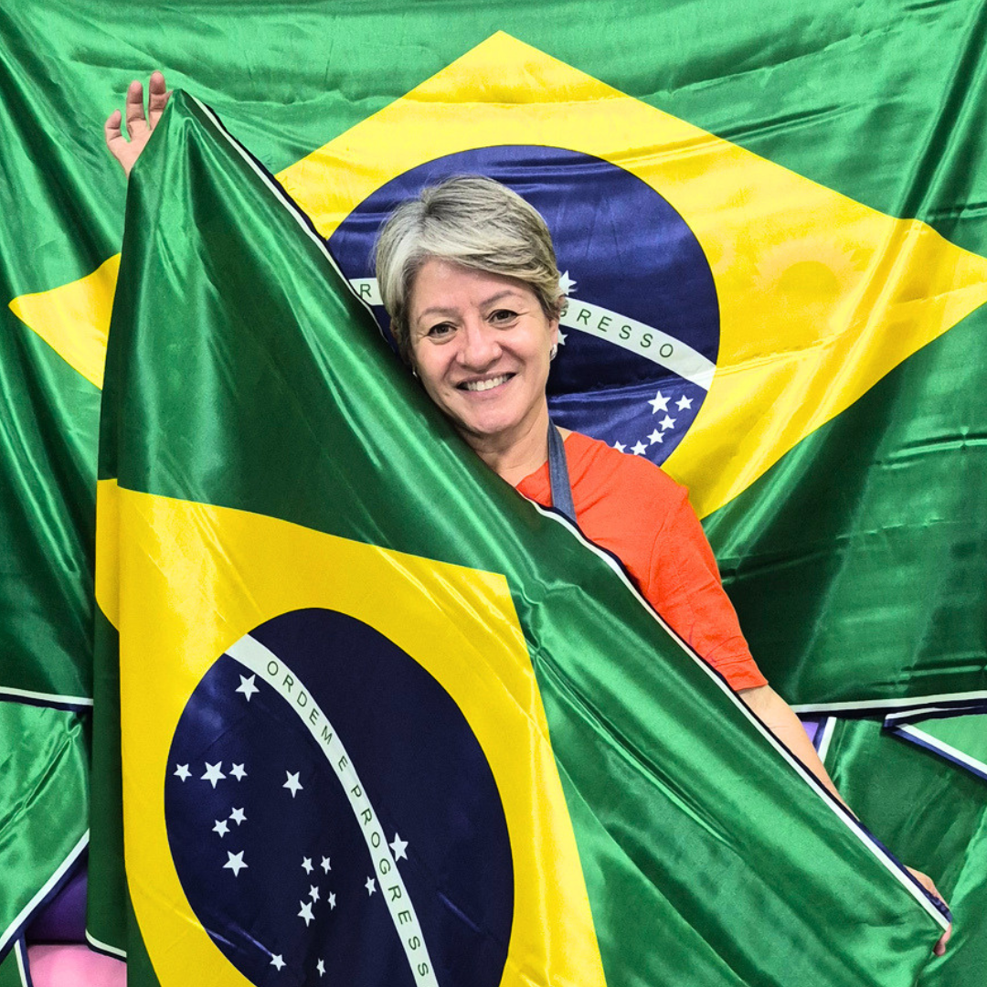 Adriana, da Adriana Armarinhos, sorrindo com uma bandeira do Brasil em meio a rolos de tecido na loja